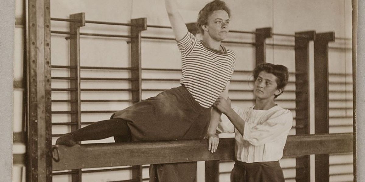 A woman balancing on a beam, with her leg and arm in the air. Another woman holds her arm to support her.