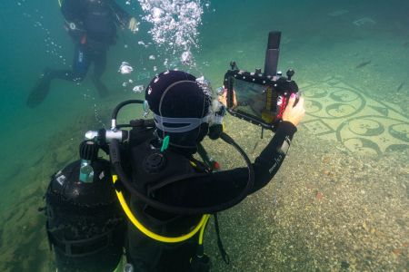 Swimming diver holds underwater tablet and points it towards a mosaic floor on the seabed.