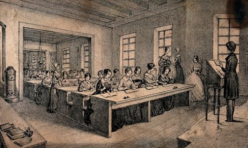 A classroom with children sitting at long tables and a teacher standing with a book in her hand
