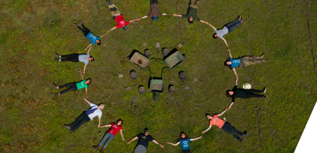 An aerial view of 14 people lying down on grass, hand in hand, forming a circle. In the centre of the circle are archaeology tools - wheel barrows, buckets, spades and trowels