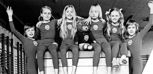 A team of young gymnasts sit arm in arm on top of a gymnastics vaulting box
