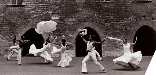 Seven Black dancers in white costumes that catch the air dance in front of a stone wall with three dark arches.
