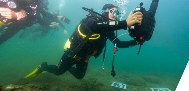 A swimming diver holds an underwater tablet. Two QR codes lie on the seabed.