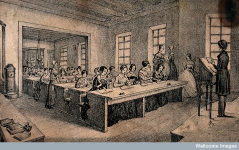 A classroom with children sitting at long tables and a teacher standing with a book in her hand - J.B. Sonde,The Wellcome Library, CC-BY