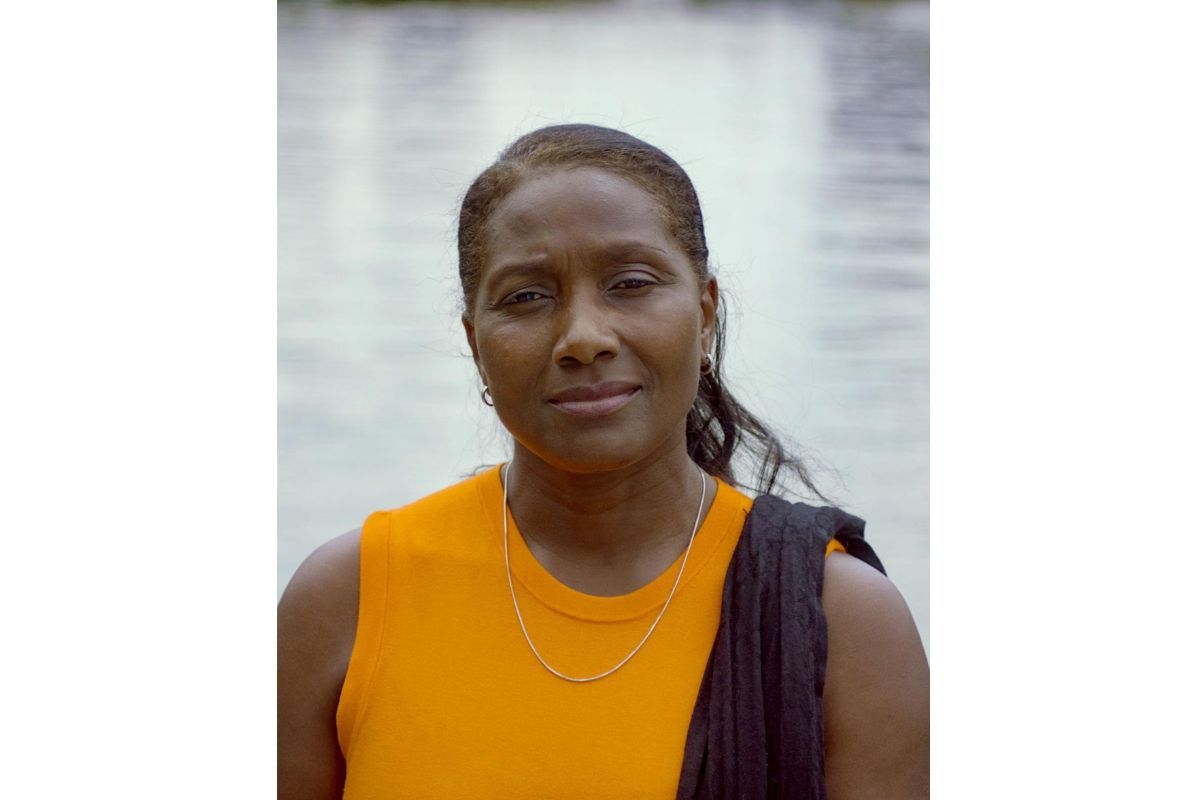 Photograph portrait of Maureen Mollis, wearing an orange vest with a black sash, and a gold chain necklace, standing in front of a body of water. 