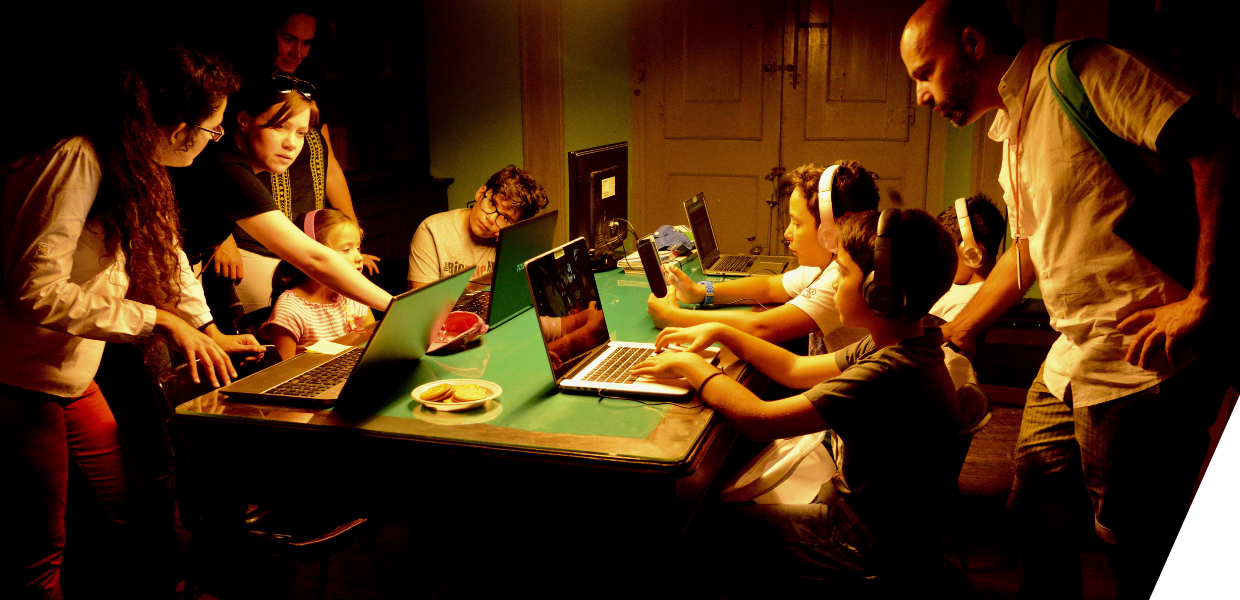 Children and adults around a table with multiple laptops and devices
