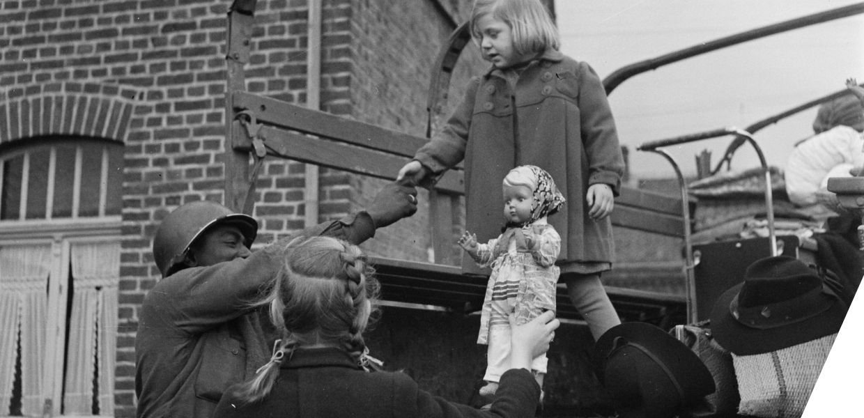 A Black soldier from the 3454th Quartermaster Truck Company helps two children climb down from the back of a truck. One child carries a large doll.