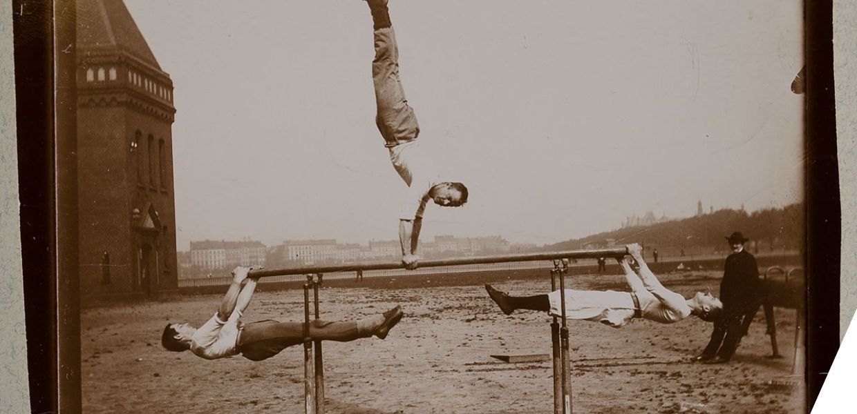 Men balancing on a beam, two holding on underneath each end; one doing a handstand in the middle of the beam. A man in a dark coat and hat looks on. 