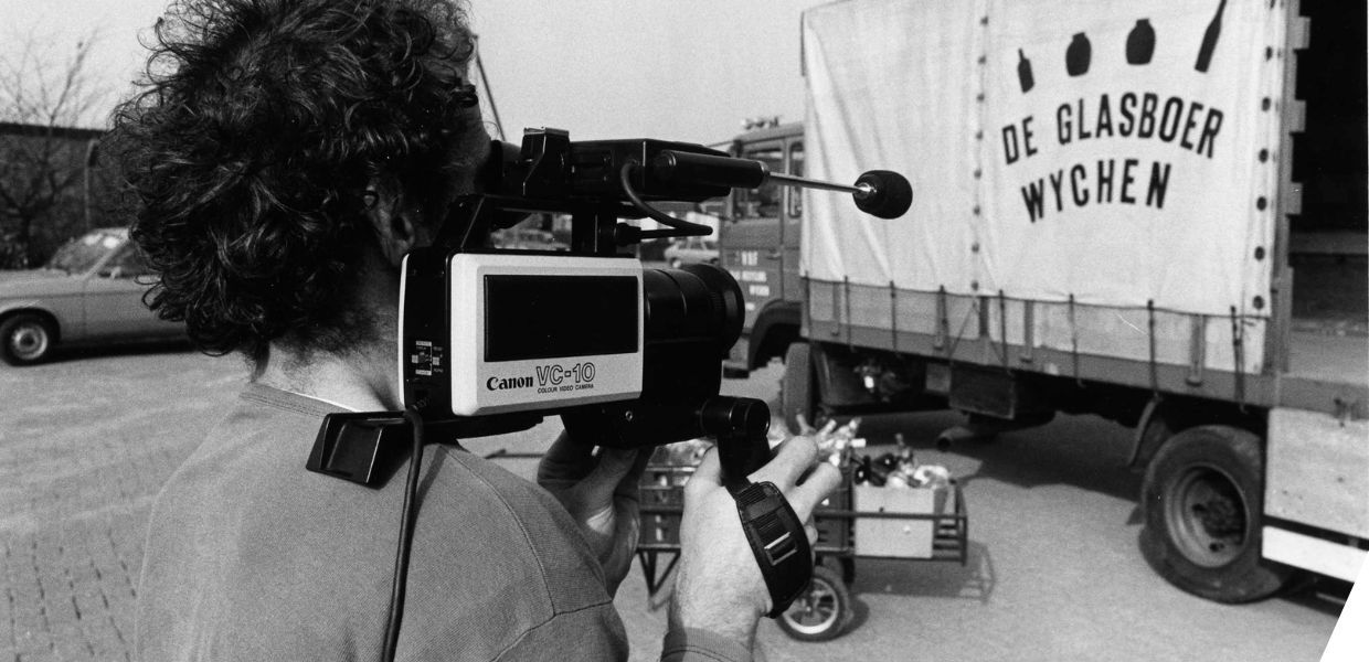 A person operating a handheld camera points it at a lorry which has the words 'De glasboer wychen' on the side