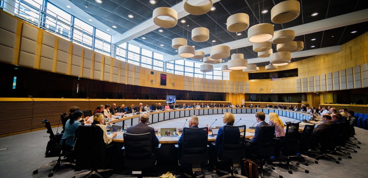 Participants at the High-level event of the Twin it! 3D for Europe s culture campaign are seated around a large curved table, in a modern, paneled room with large lamps hanging from the ceiling