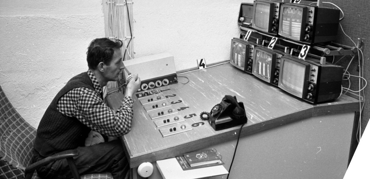 A man seated at a table. He speaks into a microphone while watching several TV screens.